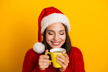 Smiling girl wearing santa hat and red sweater enjoys warm coffee mug against bright yellow background in festive winter mood