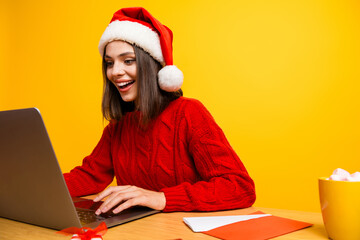 Humorous holiday scene as a cheerful young woman in a red sweater and Santa hat enjoys a laptop session amidst festive decor yellow backdrop