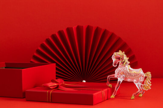 An elegant transparent horse figurine with gold accents on a bright red background with traditional fans and gift boxes. Happy Chinese New Year and the upcoming Year of the Fire Horse 2026.