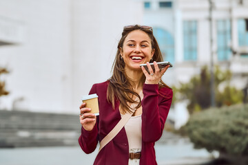 Smiling woman in burgundy blazer sending a voice message while exploring the city and holding coffee cup to go.