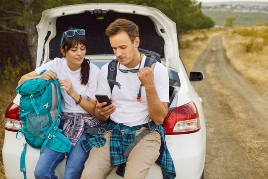 Couple by car use phone, map, backpack. Stranded on a rural road, travelers check geolocation, seek signal, and study a map while resting on the trunk near the forest. Concept: lost on the road.