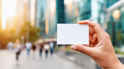 Hand holding blank business card in urban setting with blurred people walking in background, showcasing professional networking opportunities and branding