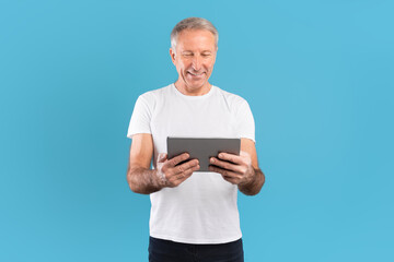 Confident mature man stands alone in front of a blue wall, smiling brightly while holding a tablet. He is engaged in watching content and appears cheerful and positive.