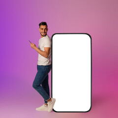 A joyful young Arab man interacts with his mobile device while leaning against a large smartphone...