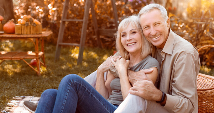 A joyful couple sits together in their backyard, surrounded by autumn leaves and pumpkins. They share a warm embrace and smile at each other, enjoying the cozy atmosphere of the season.