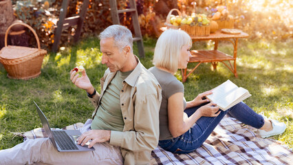 A man is focused on his laptop while eating a snack, and a woman is reading a book beside him. They are seated on a blanket in a sunny garden surrounded by greenery.