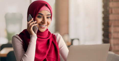 A woman wearing a red hijab smiles while on a phone call. She is seated at a table in a modern cafe, using her laptop, indicating a productive atmosphere filled with focus.