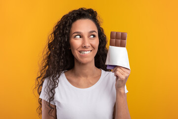 A funny hungry lady with curly hair is holding a chocolate bar and biting her lip in excitement. She stands in front of a bright yellow-orange wall, showcasing her love for sweet treats.