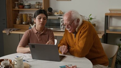 Front view shot of Latin American young woman as social worker helping senior man using laptop and working with bills and taxes at home sitting at table together