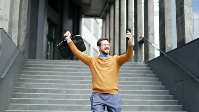 Happy excited businessman celebrating success while receiving great news on phone walking down stairs outside office building. Smiling shocked surprised entrepreneur cheerfully rejoices in achievement
