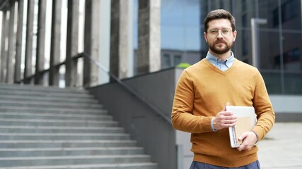 Portrait of serious male teacher or professor holding books laptop looking at camera standing near university building. Confident scientist. Empty copy space for writing text. Template for advertising