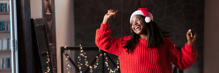 A cheerful young woman celebrates the holiday season by dancing in a cozy room adorned with twinkling lights. She wears a bright red sweater and a Santa hat, exuding joy and festivity.