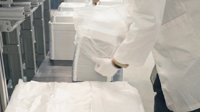 Close up hands of worker in white coat and gloves packing stack of aluminum containers into transparent plastic bag. Industrial packaging process in clean manufacturing environment.