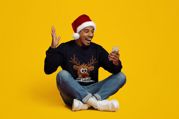 A joyful man with a Santa hat sits cross-legged on the floor. He is smiling and waving while looking at his smartphone. The bright yellow background adds a festive touch to the scene.