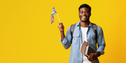 A young man stands against a vibrant yellow backdrop, smiling widely. He holds a small British flag...