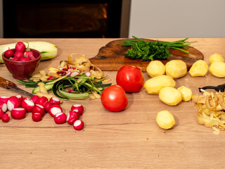 Fresh vegetables and kitchen scraps on a wooden countertop during meal preparation. Radishes, potatoes, cucumber, tomatoes and fresh herbs.