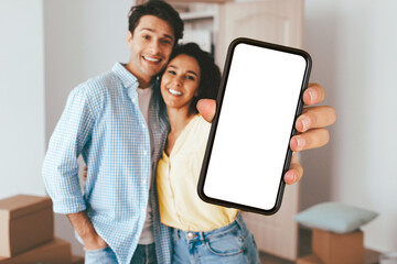 A cheerful couple stands close together, smiling happily while holding a blank smartphone. Behind them, moving boxes are scattered, indicating they just moved into a new home.