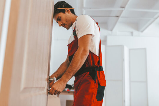 A dedicated worker in red overalls carefully installs door hardware in a contemporary room. The scene emphasizes skill and attention to detail in home improvement.