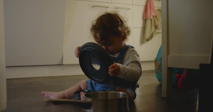 Toddler sitting on kitchen floor exploring pot and lid with curious focus, holding cookware and studying shapes during quiet independent play at home
