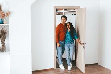 A young couple stands at the doorway of their new home, smiling and excited. The bright room has neutral colors and a cozy feel. They are ready to start this new chapter together.