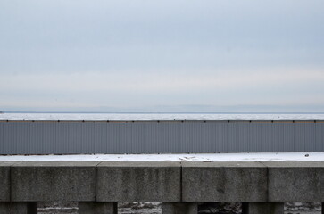 A frozen sea. The Gulf of Finland is covered in ice. Snow lies on the ice in places, and ice is visible. People are walking on the ice in the distance. The beach is fenced with a gray metal fence.
