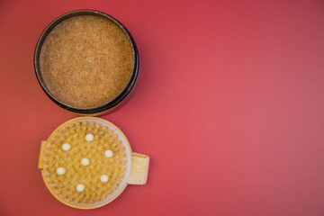 Body scrub in black container next to exfoliating brush on pink background, highlighting essential skincare products for a rejuvenating beauty experience