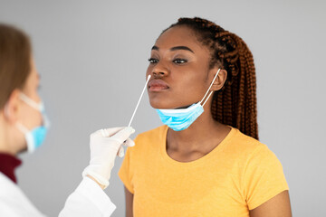 A millennial black woman receives a nasal covid-19 swab test from a doctor wearing gloves. The setting is a grey studio background, focusing on viral disease protection.