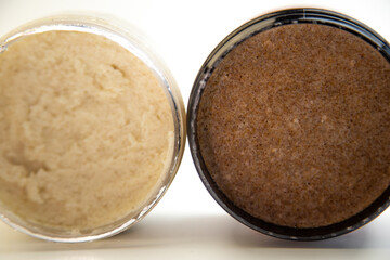 Two jars containing natural body scrubs, one light beige and the other dark brown, highlighting unique textures and colors, perfect for beauty and skincare applications