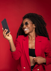 Smiling woman in red blazer and sunglasses taking selfie with smartphone on red studio background