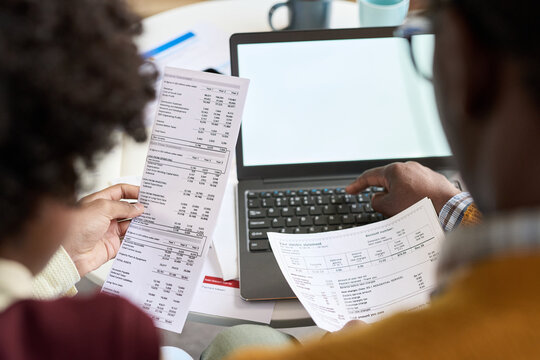 Black man and young child sitting together analyzing financial documents and using laptop, hands holding printed statements