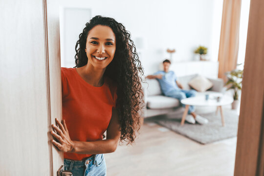 A woman with curly hair stands at the door, smiling warmly at the camera. In the background, a man is seated comfortably on a couch, creating a relaxed home atmosphere.
