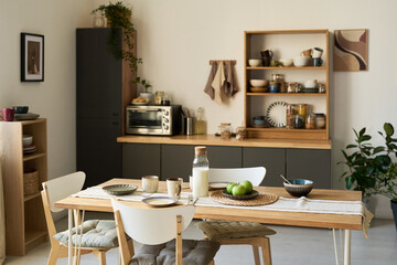 Modern kitchen interior featuring wooden dining table set with plates, bowls, glass of milk, green apples, and carafe, open shelving displaying dishes and plants in background