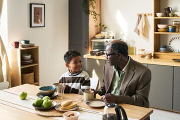 Senior man sitting at table talking with Black child boy in kitchen, both smiling and making eye contact while sharing breakfast, food and drinks on table