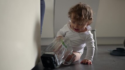 Curious toddler on kitchen floor exploring empty blender jar with playful concentration and joyful wonder during morning moment filled with innocence and simple discovery