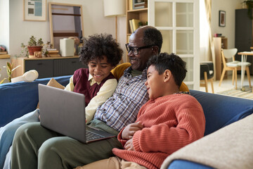 Senior Black man sitting on sofa with two children using laptop, smiling and interacting together, multi generational family bonding in living room, children appearing engaged