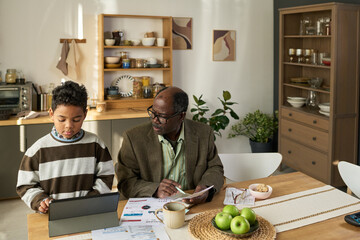 Boy using tablet at kitchen table while senior Black man sitting beside him holding documents and pen, both focused on paperwork and technology in home setting