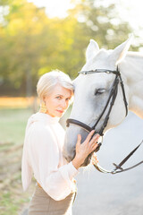 Obraz premium An iconic, beautiful woman standing and holding a white horse in a park at sunset and love horse