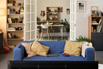 Empty living room with blue sofa positioned in foreground, remote control, laptop, and game controller resting on cushions, open double doors revealing dining area in background