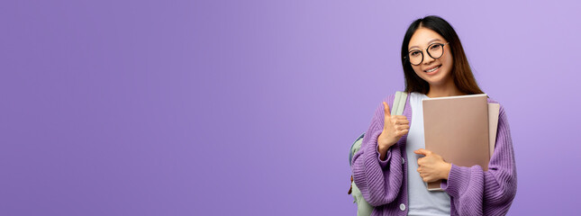 A young woman with long hair and glasses stands confidently against a purple background. She is smiling and giving a thumbs up while holding a stack of books and a backpack.
