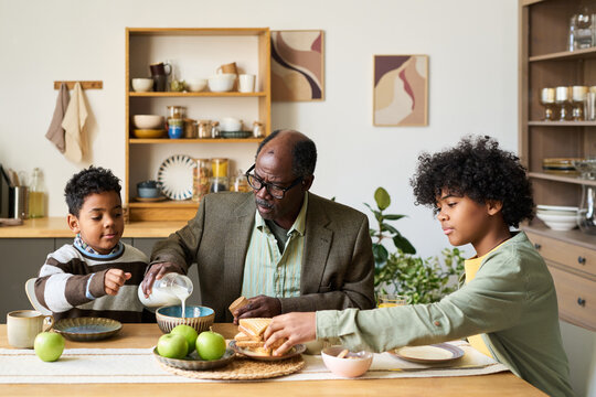 Senior Black man sitting at kitchen table with two Black boys, pouring milk into bowl while boys preparing breakfast, family members interacting during morning meal at home
