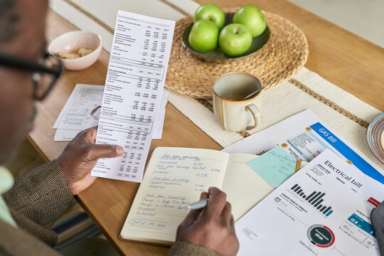 Mature Black man reviewing utility bills and writing expenses in notebook at kitchen table, holding paper with financial data, surrounded by documents and green apples