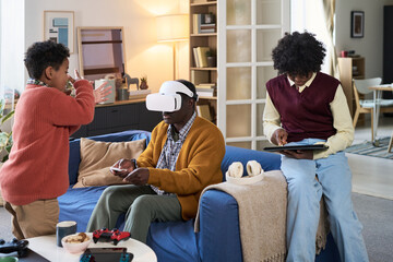 Black mature man wearing virtual reality headset sitting on sofa interacting with game controller while Black teenage boy using digital tablet