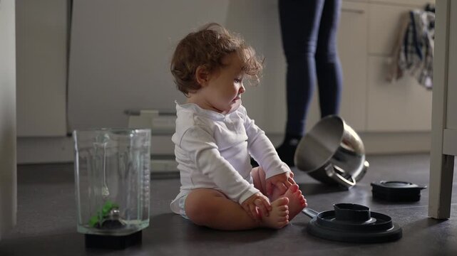 Curious toddler sitting on kitchen floor surrounded by scattered cookware while studying blender parts with calm focus during gentle family moment filled with early exploration at home