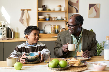 Boy sitting at kitchen table eating breakfast with senior Black man drinking from mug and smiling, both engaging in conversation, kitchen shelves and fruit in background
