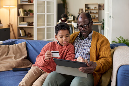 Child holding credit card sitting with Black senior man using digital tablet together on sofa, both focusing on screen, intergenerational family spending time with technology