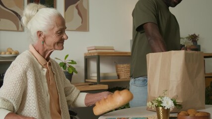 Tilt up shot of senior woman using wheelchair at home and watching social worker unpacking groceries and food delivery on kitchen table, copy space - Powered by Adobe