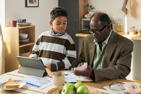 Black senior man reviewing bills and documents at table while Black boy standing beside him using digital tablet, both focused on paperwork in home kitchen setting, financial planning scene
