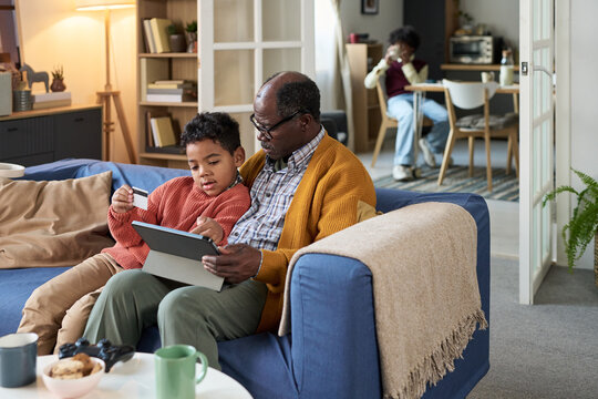 Senior man sitting on sofa helping Black boy using digital tablet in modern living room