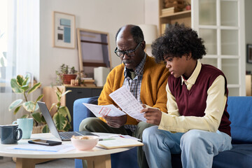 Mature Black man and teenage Black boy sitting together reviewing financial documents, using laptop on table, focused on paperwork, collaborating on home budget planning