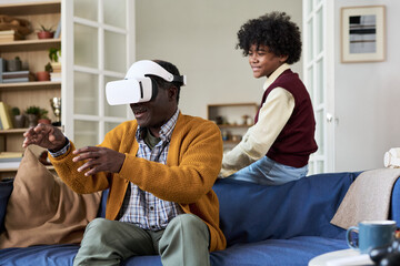 Mature Black man wearing virtual reality headset interacting with digital environment while Black teenager sitting on couch in background observing and smiling